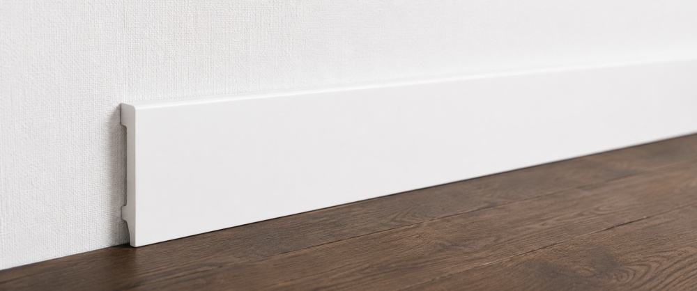 Witte kunststof plint tegen witte wand met donkere houten vloer